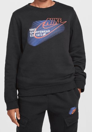 Personne portant un sweat-shirt et un pantalon noirs Nike avec un logo et un texte Nike bleus et orange "Nike Sportswear Est. 1972" sur le sweat-shirt et le pantalon.