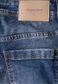 Blauer Denim-Stoff mit einer strukturierten Oberfläche, versehen mit gelben Nähten und einem beigen Lederetikett, das mit "WE LOVE DENIM" und "CECIL" geprägt ist.