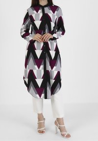 Robe chemise longue colorée avec un motif géométrique en violet, gris et noir, dotée d'une fermeture à boutons sur le devant et d'un ourlet asymétrique, portée avec un pantalon blanc.