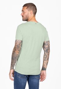 Camiseta de manga corta verde claro con un escote redondeado y una textura suave, combinada con jeans azules, mostrando intrincados tatuajes en los brazos.