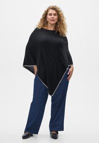 Črni pončo trikotne oblike, z teksturiranim robom in kontrastnim svetlim obrobjem, nosi čez modre široke hlače.