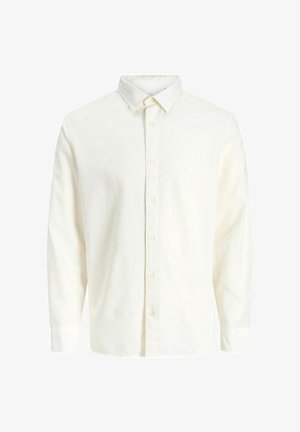 Garçon portant une veste bomber beige, une chemise blanche, un pantalon kaki, des chaussures marron et une casquette blanche, debout et souriant devant un fond clair uni.