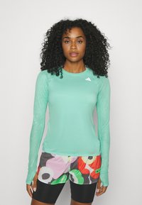 Mintgrön långärmad tränings tröja med texturerade ärmar och rundad halsringning, parat med mönstrade, färgglada shorts och svarta cykelbyxor.