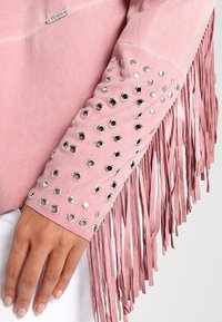 Roze suède jasje met een mouw die zilveren grommets en lange franjes heeft. Soepele textuur met een slank ontwerp en subtiele branding.