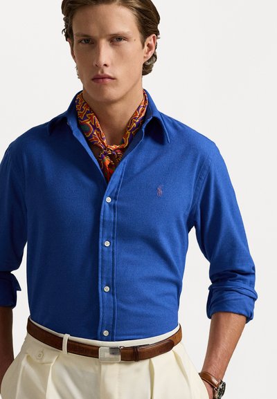 Blue long-sleeve shirt with a buttoned front, collar, and a small logo. Paired with a multicolored patterned scarf and beige trousers.