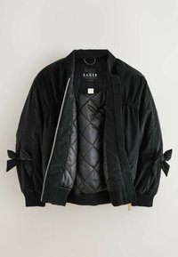 Giacca bomber nera in nylon trapuntato con polsini a costine. Presenta fiocchi sulle maniche e una chiusura con zip. Interno foderato in tessuto morbido.