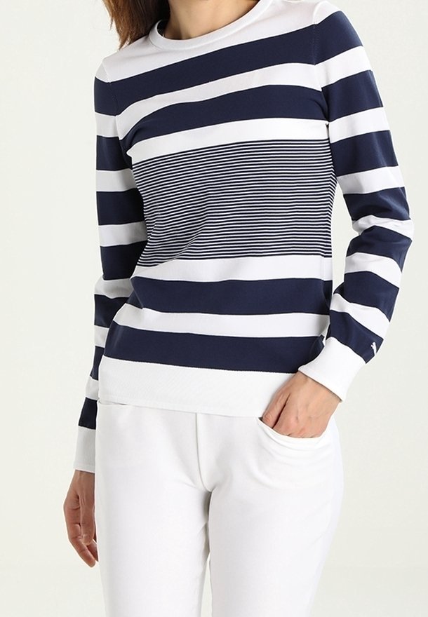 Femme portant un chemisier à manches longues à rayures marines et blanches avec des rayures horizontales étroites au centre, associée à un pantalon blanc.