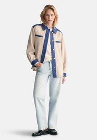 Giacca in camicia bicolore beige e blu con maniche lunghe, chiusura a bottoni e due tasche sul petto. Indossata sopra un top crema e jeans in denim chiaro.