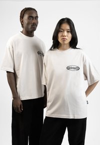 Beige cotton oversized t-shirts feature a black graphic on the chest. Paired with black corduroy pants, they contrast with the light tops.