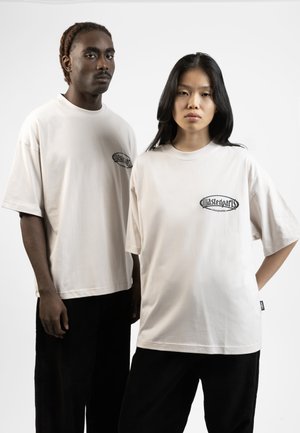 Des t-shirts oversize en coton beige présentent un graphique noir sur la poitrine. Associés à des pantalons en velours côtelé noir, ils créent un contraste avec les tops légers.