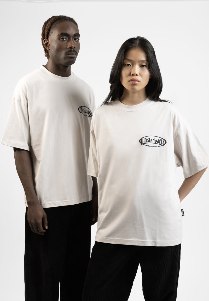 Beige cotton oversized t-shirts feature a black graphic on the chest. Paired with black corduroy pants, they contrast with the light tops.
