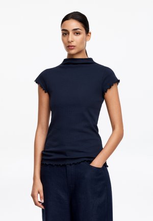 Femme aux cheveux foncés attachés, portant un haut court bleu marine ajusté et un pantalon bleu marine, une main dans la poche.
