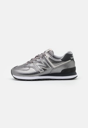 Sølvfarvet New Balance sneaker med grå snørebånd, hvid mellemsål og sort ydersål, vist fra siden på en hvid baggrund.