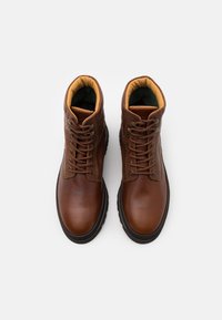 Bottes de cheville en cuir marron avec des accents texturés, bout rond et semelles en caoutchouc noires. Devant lacé et col rembourré en tissu beige.