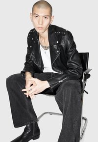 Jachetă de biker din piele neagră cu ținte argintii, tricou alb și pantaloni din denim închis. Poziție așezată într-un scaun modern, purtând cizme negre până la gleznă.