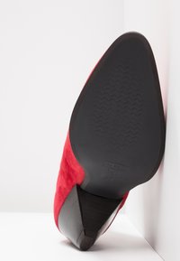 Chaussure à talon haut en daim rouge avec bout pointu, semelle noire à surface antidérapante, photographiée par dessous sur fond blanc.