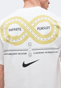 Πίσω μέρος λευκού πουκάμισου με χρυσό ατελείωτο αγωνιστικό κύκλωμα με την ετικέτα "INFINITE PURSUIT", ενθαρρυντικό κείμενο και μαύρο λογότυπο Nike swoosh κάτω.