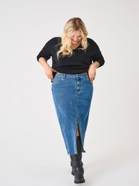 Blonde Frau, die einen schwarzen Pullover, einen blauen Denim-Midirock mit Frontschlitz und schwarze kniehohe Stiefel trägt, schaut nach unten und lächelt.