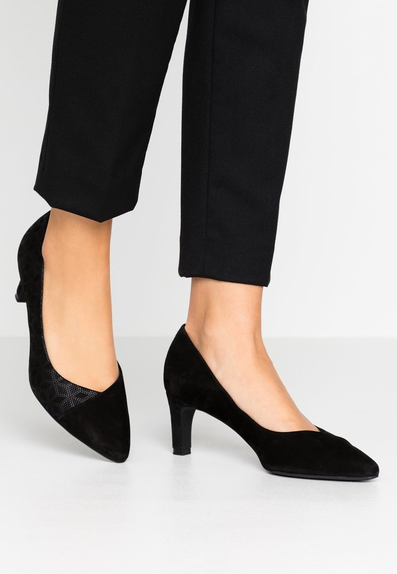 Chaussures noires à talon moyen et bout pointu portées avec un pantalon noir raccourci sur un sol blanc, montrant les chevilles et les pieds de côté.