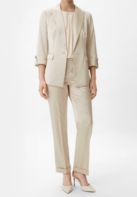 Kvinde iført en beige skræddersyet blazer med opbrettede ærmer, matchende højtaljede bukser og beige spidse højhælede sko.