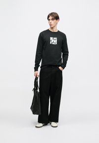 Μαύρο μπλουζάκι με μακριά μανίκια που διαθέτει γραφικό λογότυπο, συνδυασμένο με φαρδύ μαύρο παντελόνι και λευκά αθλητικά παπούτσια. Το μοντέλο κρατά μια μαύρη τσάντα.