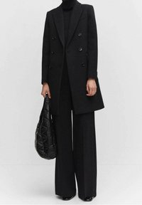 Sort dobbeltradet frakke med skrå lapels, parret med sorte brede bukser. Holder en tekstureret sort taske, alt sammen i et minimalistisk design.