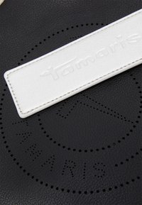 Prodotto in pelle nera con etichetta bianca in rilievo con la scritta "Tamaris", motivo di fori circolari e texture liscia.