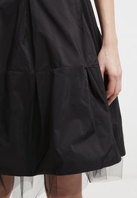 Jupe noire à design en couches, avec un tissu lisse et un ourlet en tulle. Plis et mise en forme structurée sont visibles, mettant en valeur sa silhouette.