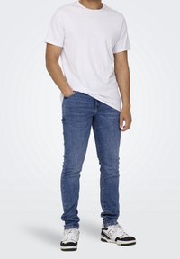 Valkoinen lyhythihainen t-paita, siniset tiukat farkut ja mustat/valkoiset tennarit. Sujuva kangas, istuva muotoilu. Farkuissa on klassinen viiden taskun tyyli.