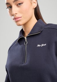 Tummansininen vetoketjullinen huppari, jossa on korkea kaulus, ja jossa on valkoisella brodeerattu "New York" -teksti sekä hopeinen vetoketjuyksityiskohta. Pehmeä pinta.