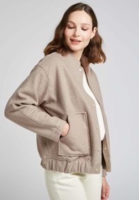 Veste marron clair en tissu texturé, avec un col rond, des boutons-pression et des poches latérales. L'ourlet volanté en bas ajoute une touche de détail.