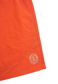 Helle orangefarbene Shorts aus leichtem Stoff, mit einer abgerundeten Tasche und einem weißen Logo-Patch mit Text am unteren Saum.