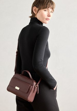 Kobieta ubrana w czarny golf i spodnie, niosąca na ramieniu teksturowaną ciemnobrązową skórzaną torebkę na pasku, spoglądająca przez ramię.