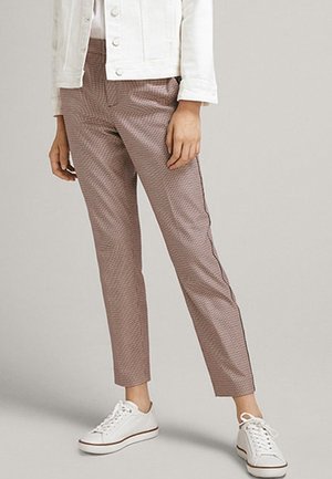 Person trägt schlanke, knöchellange, braun gemusterte Hose, weiße Turnschuhe und eine weiße, geknöpfte Jacke und steht vor einem schlichten Hintergrund.