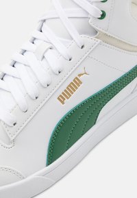 Bílé kožené tenisky s zeleným akcentovým pruhem, zlatým logem "PUMA" a bílými tkaničkami. Texturovaná podrážka a hladké povrchové detaily.