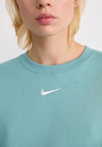 Primer plano de una persona que lleva un sudadera de color azul claro-verde con un logo de swoosh blanco de Nike en el pecho.