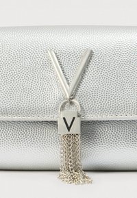 Srebrna torbica od umjetne kože s teksturiranom površinom, s istaknutim srebrnim logom "V" i detaljem s lancem u obliku resice.