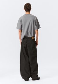 Šedé tričko s krátkým rukávem v kombinaci s volnými tmavými džíny. Mezi výrazné detaily patří nášivka na pase a boční kapsy.