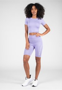 Ljubičasti kratkotrajni crop top s kratkim rukavima i biciklističke kratke hlače visokog struka s "GORILLA WEAR" logotipom. Glatka, rastezljiva tkanina, uparena s bijelim sportskim cipelama.