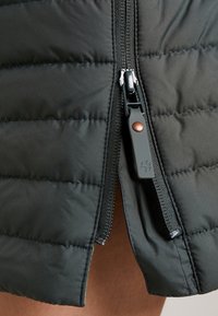 Detailní záběr na černou prošívanou bundu, zip je částečně rozepnutý, ukazuje gumovou táhlo s malým designem tlapky.