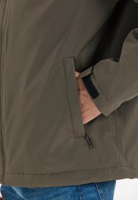 Oliven grøn overtøjsjakke med en glat tekstur. Funktioner inkluderer en sidelomme, justerbare manchetter med Velcro og en skræddersyet pasform.