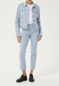 Veste en denim bleu clair avec pantalon assorti bleu clair. Sous-vêtement blanc à col rond. Mocassins noirs complètent la tenue.