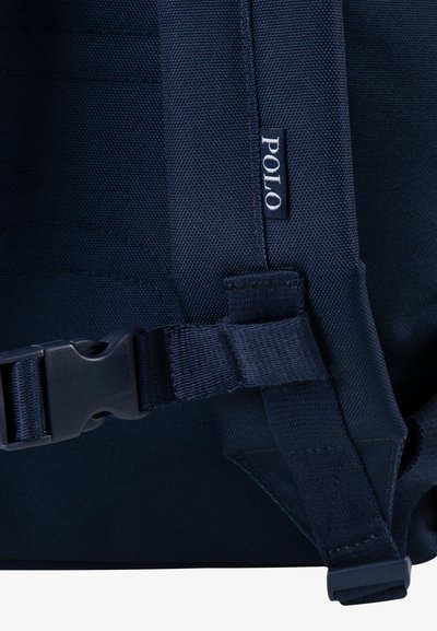 Navy modra nahrbtnikova zanka, ki je narejena iz trpežne tkanine, ima plastično zaponko in našit logotip na strani, kar poudarja uporabnost in oblikovanje.
