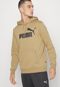 Felpa con cappuccio beige con tasca a canguro, caratterizzata da un logo Puma nero sul petto e polsini a costine. Design semplice e confortevole.