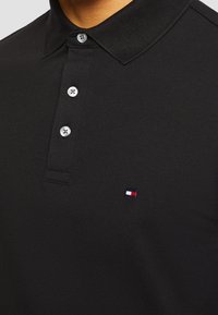 Polo noir avec trois boutons blancs et un petit logo rectangulaire rouge, blanc et bleu sur la poitrine gauche.
