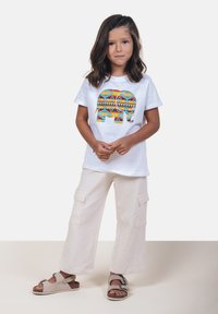Camiseta blanca con un gráfico colorido de elefante, pantalones cargo crema con bolsillos, y sandalias grises con hebillas. Diseño simple y casual.