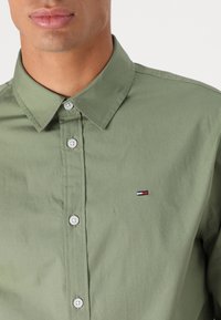 Groen overhemd met knopen, gemaakt van gladde stof; heeft een kraag en een klein geborduurd logo in rood, wit en blauw op de linkerborst.