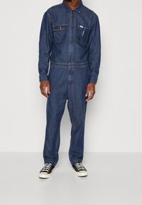 Combinaison en denim bleu foncé à manches longues avec deux poches poitrine. Comprend une fermeture à boutons, une coupe droite des jambes et des manches retroussées.