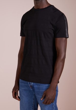 Czarny t-shirt z krótkim rękawem z subtelnymi poziomymi paskami i taśmą z logo na rękawie, noszony przez osobę w niebieskich dżinsach na jednolitym tle.