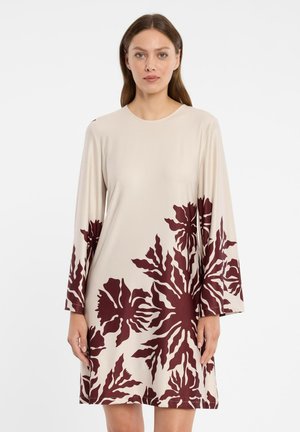 Vrouw met lang bruin haar, gekleed in een beige jurk met lange mouwen, voorzien van grote donkerrode bloemmotieven op het onderste gedeelte en de mouwen.